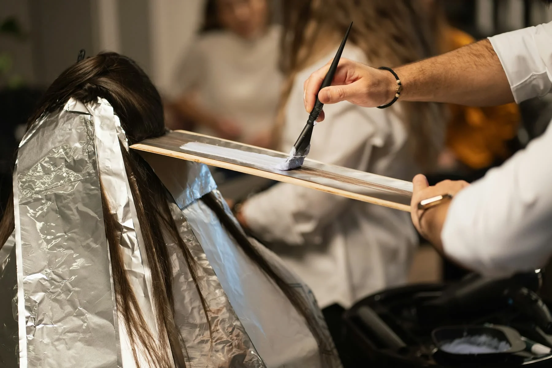 A colorist paints lightener onto a section of long dark hair over a board, with foils below